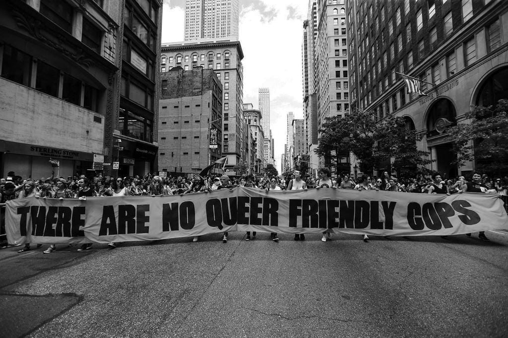 Activists carry a large banner that spans the width of the road they are marching down. It reads: THERE ARE NO QUEER FRIENDLY COPS.