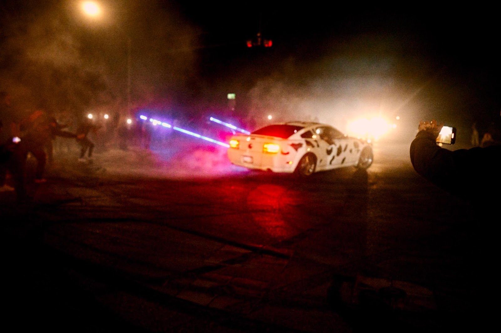 A different angle of people gathered around a car in an intersection. Tire skid marks are visible. There are some very bright flashlights pointed at the car.