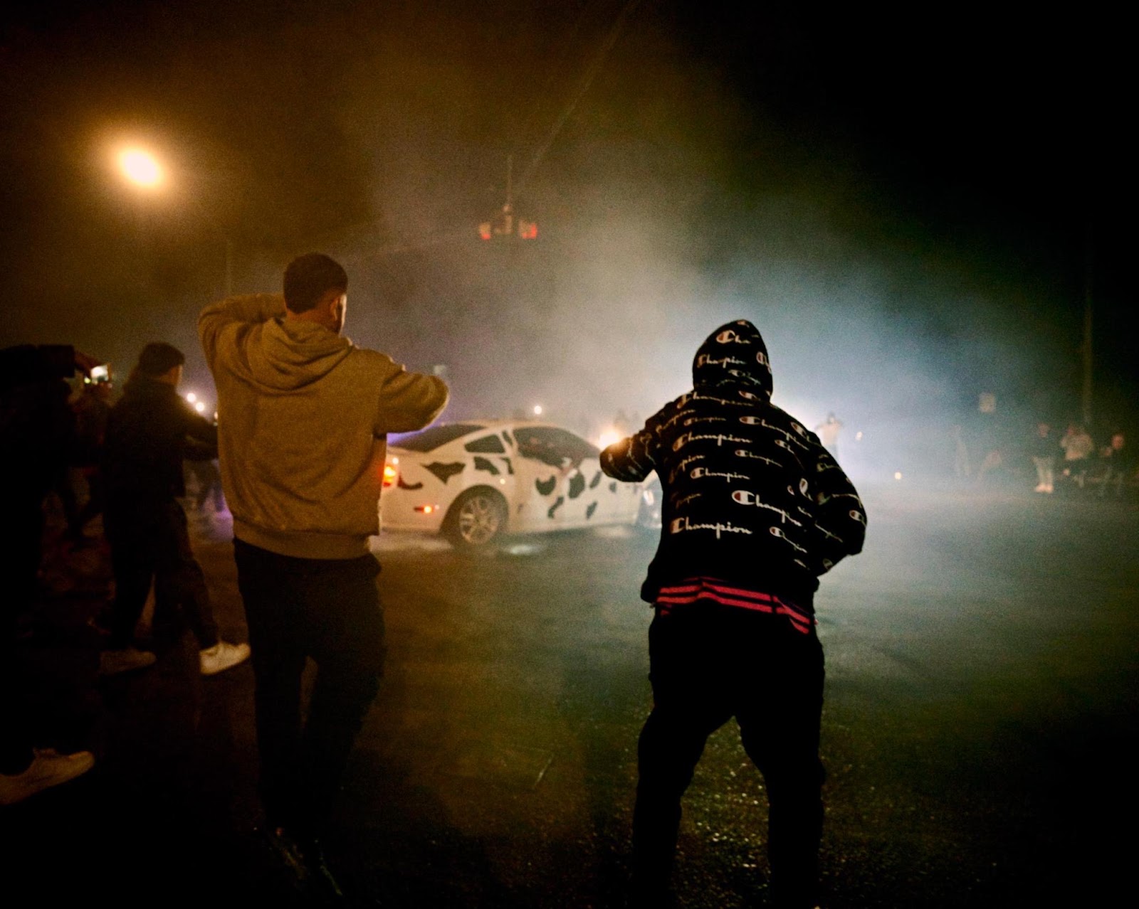 People gathered around a car in an intersection, some filming with their phones. There's smoke in the air, seemingly from the car doing donuts.