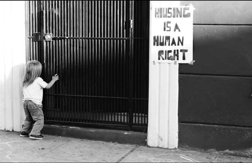 A child reaching out to a gate; nearby is a sign that reads "HOUSING IS A HUMAN RIGHT"