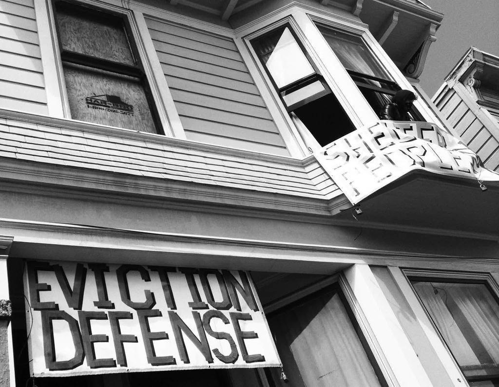 Squat with an "EVICTION DEFENSE" banner, one that says "SHELTER PEOPLE". One window is boarded up, another has a person in it.