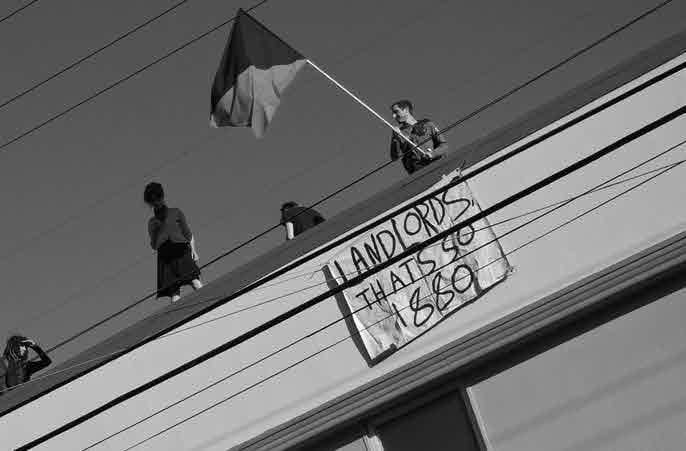 People standing on an overpass, one waving a bisected anarchist flag, with a sign dropped reading "LANDLORDS, THATS SO 1880"