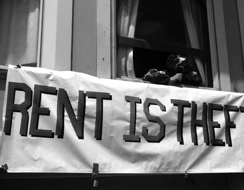 Banner reading "RENT IS THEFT" hangs under an open window with a hooded, masked sunglasses-wearing person in it