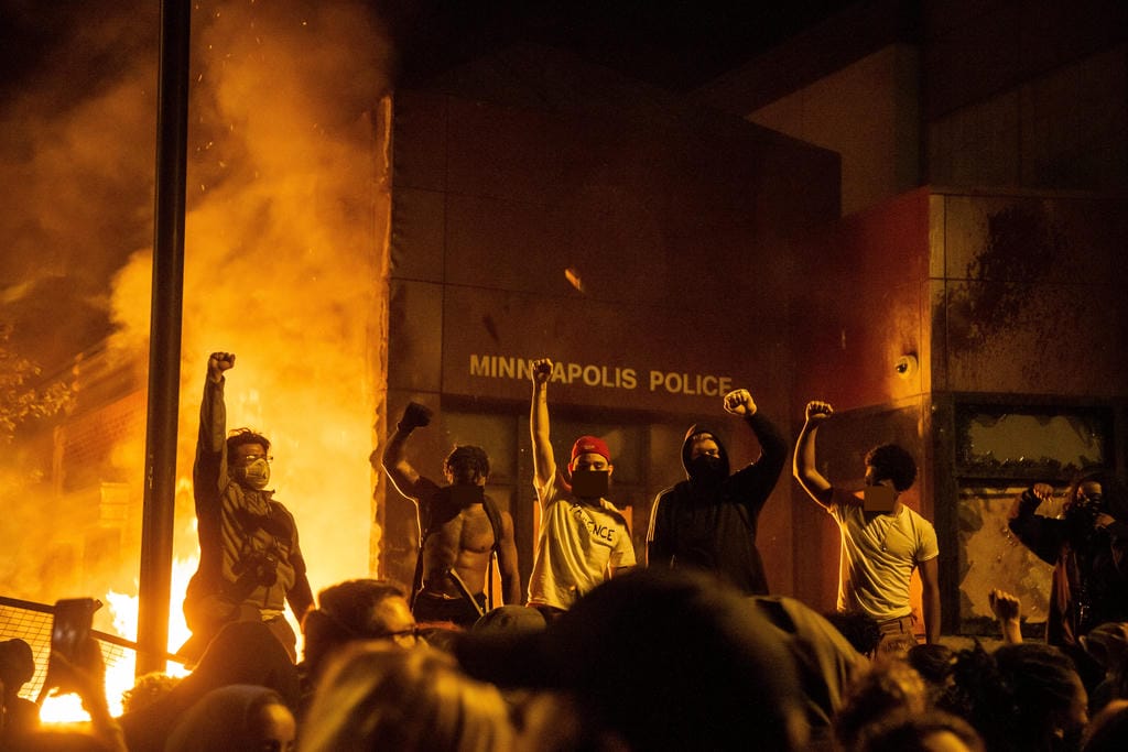 The third police precinct in Minneapolis burning during the George Floyd Uprising. Six protesters stand in front of it facing the camera with their fists raised. A large crowd faces them with their backs to the camera.