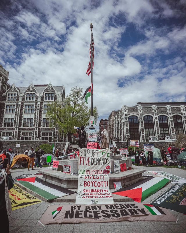 A monument in the middle of an encampment built around a flag pole, with Palestinian flags and political slogans. One prominent one says "BY ANY MEANS NECESSARY". In the center is a tall fabric sign reading "SUPPORT THE FIVE DEMANDS / VIVA PALESTINA / THE FIVE DEMANDS: 1. DIVEST! 2. BOYCOTT! 3. SOLIDARITY! 4. DEMILITARIZE! 5. A PEOPLE's CUNY!"