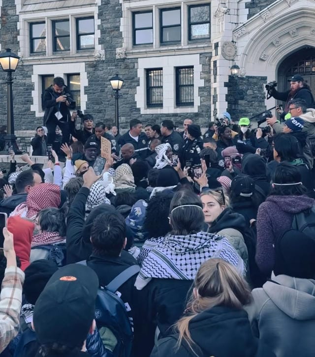 A big crowd of protesters, many in keffiyehs, jostling with police or campus authorities and several photographers.