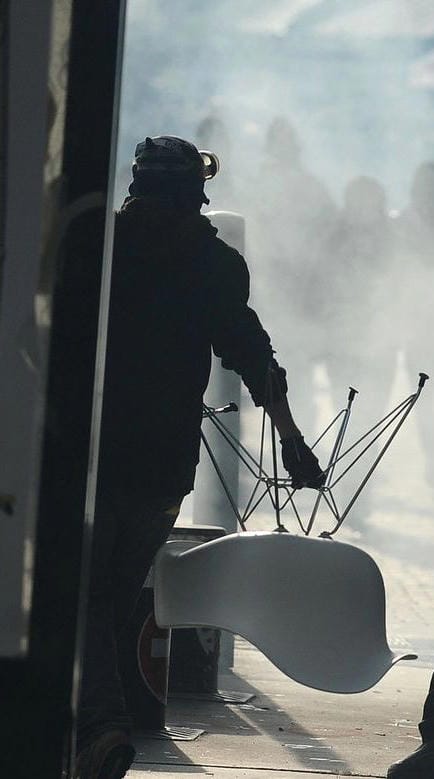A protester wearing goggles and gloves holds a fancy chair in their hand as though about to throw it. In the background we can see a concrete bollard, then further back human figures obscured by teargas or smoke.
