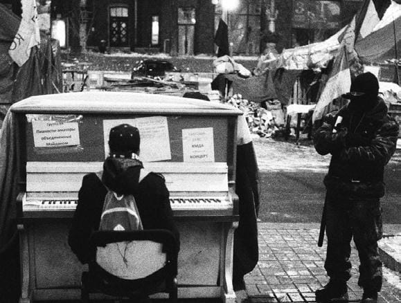 A piano outdoors with a person sitting at it. It's in some sort of encampment. A person in a ski mask and thick clothing stands to one side with a baton or similar weapon at their belt. There are various flags in the background that I can't make out that suggest the encampment is a political protest.