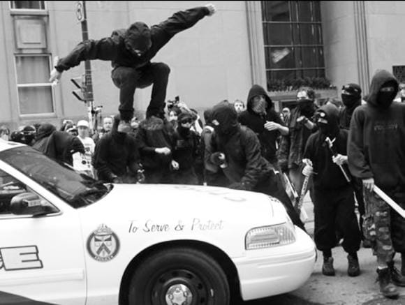 A group of people milling around a police car in bloc. One of them is jumping on the hood of the car and is caught by the photo in midair.