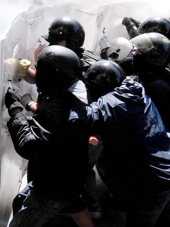 A group of activists in motorcycle helmets holding an improvised shield push against the police from the previous image.