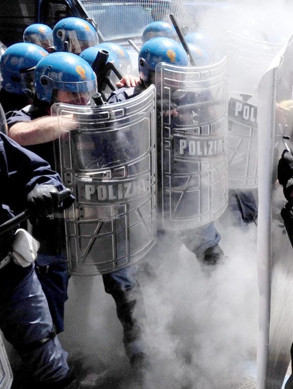 Riot police wearing blue helmets and holding shields that say POLIZIA brace themselves against activists pushing against them using an improvised shield which is barely visible. There is teargas or smoke visible. The cops have some sort of guns, maybe beanbag launchers.