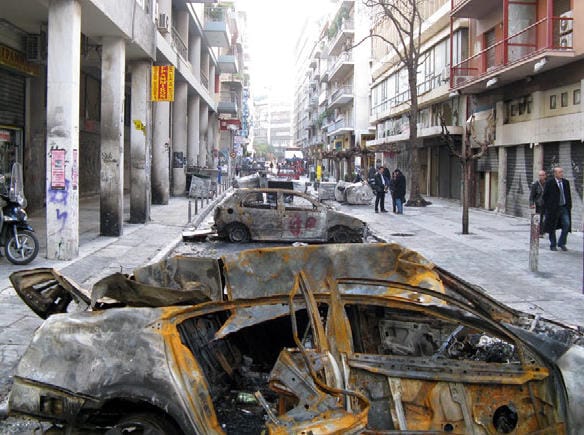 A city street with several burned out cars. There's anarchist graffiti on a nearby cement pillar. Some businesspeople are strolling by.