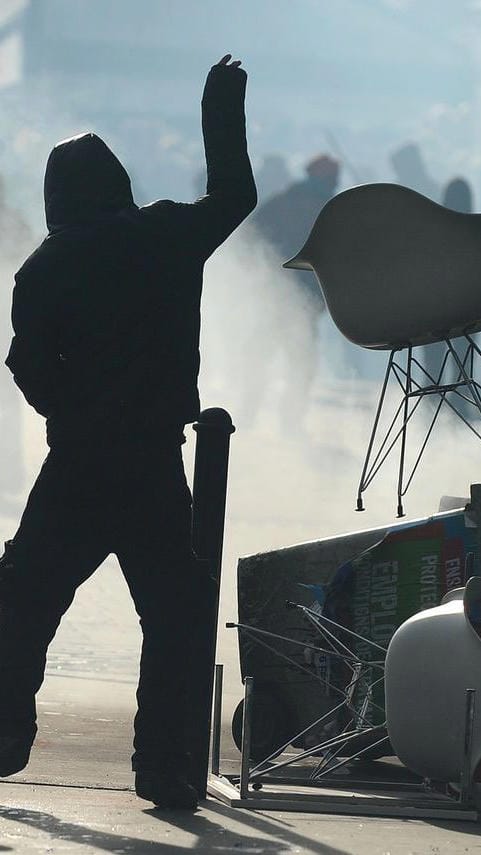 An activist, perhaps the one from the previous image, throwing a fancy chair. There's a pile of objects visible next to a metal bollard, seemingly an improvised barricade.