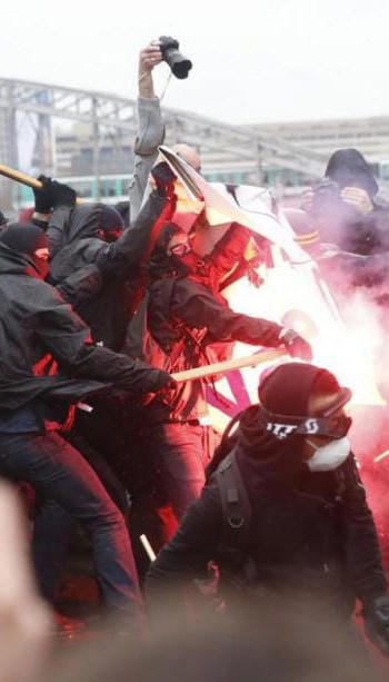 A chaotic scene of activists in bloc, prepared with shields and goggles and masks, react to a red flare. Some of them are swinging poles. There are two photographers in the back, one of whom is lifting their camera to the full reach of their arm to try to see over the crowd.
