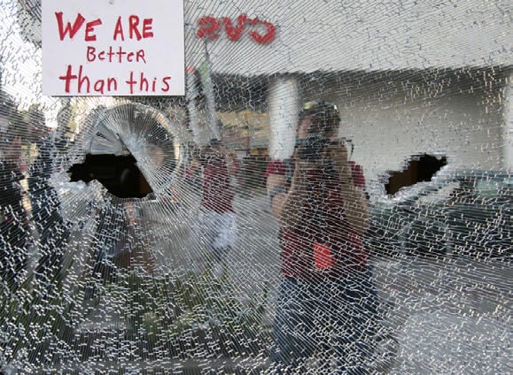 Photo of smashed glass with a handwritten note on it that reads "WE ARE BetteR than this". In the reflection of the glass a CVS storefront, car, and some people are visible, including the photographer who has one of those fancy big cameras.
